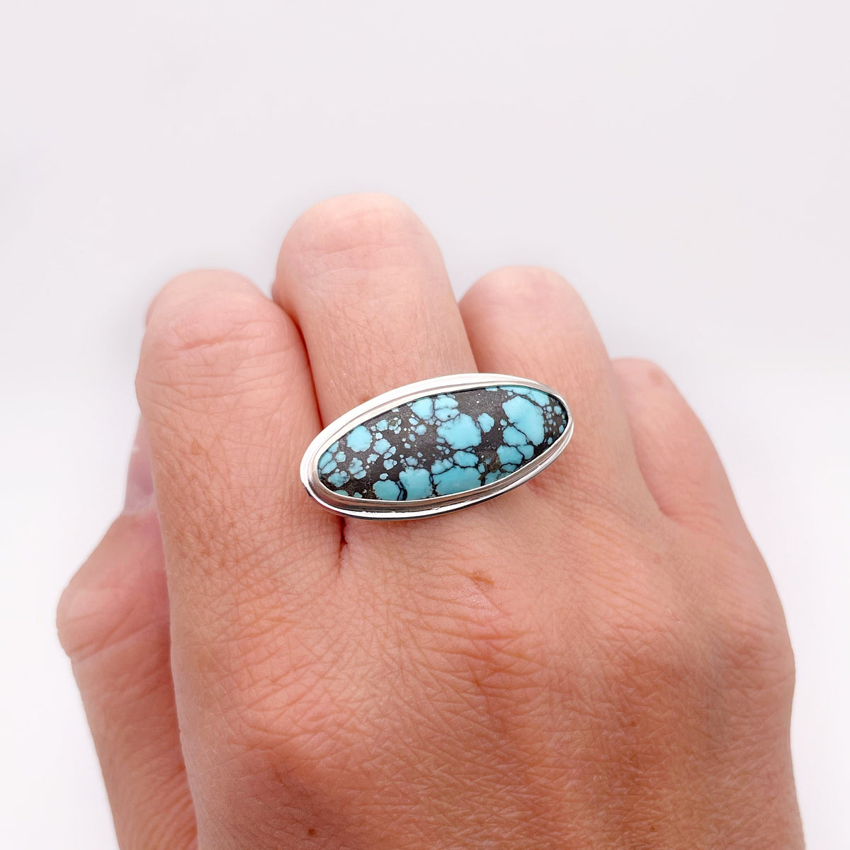 A silver ring featuring an oval turquoise stone with dark matrix, worn on a person's hand against a plain white background.