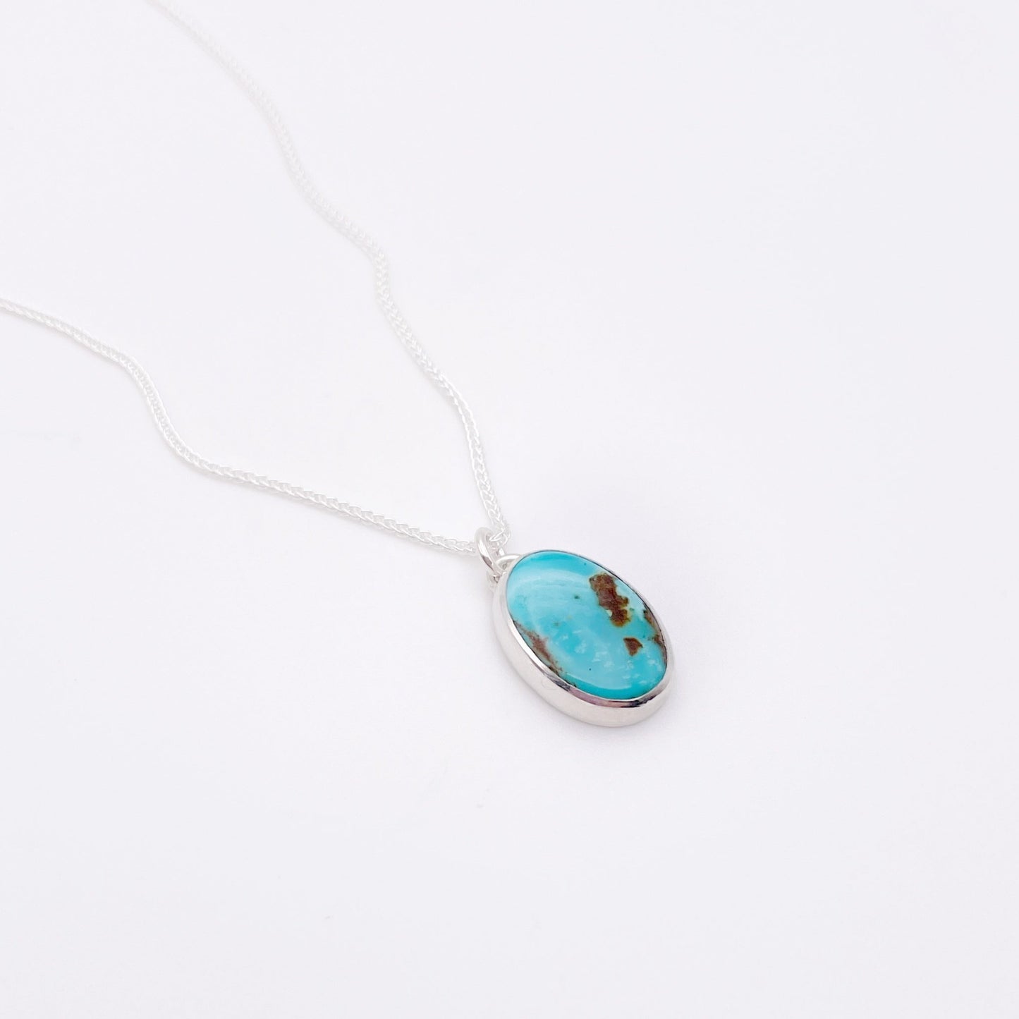 A close up detail image of vertical oval of blue green turquoise with a small bail on top and hanging from a delicate silver link chain on a white background