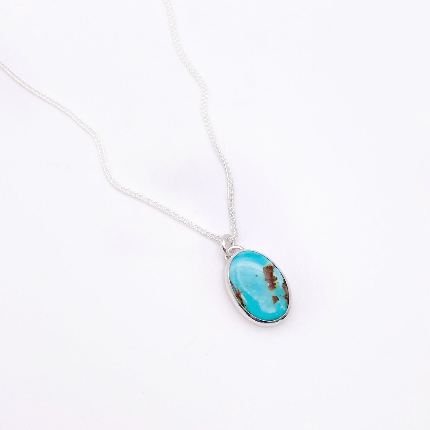 A vertical oval of blue green turquoise with a small bail on top and hanging from a delicate silver link chain on a white background